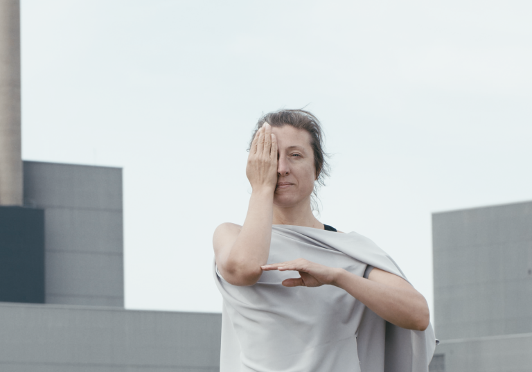 En dansare i grå bljande tunika står framför BArsebäcks kärnkraftverk. Hon håller en hand för ena ögat och skapar en vinkel med sina armar. 