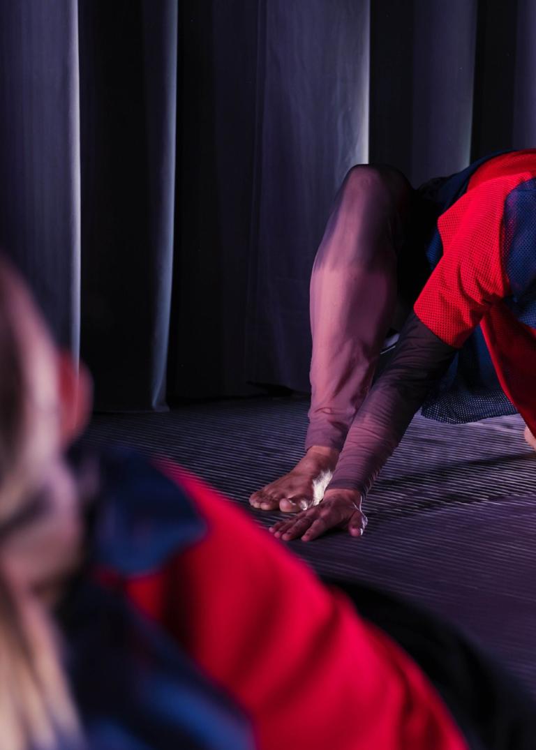 En dansare i röd och blå kläder halvsitter på knä och händer golvet och ler nyfiket mot en annan dansare i förgrunden.