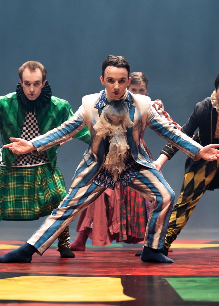 Fyra dansare i färglada hovdräkter dansar på ett färglatt golv. De böjer sig framåt med armarna öppna åt sidorna.