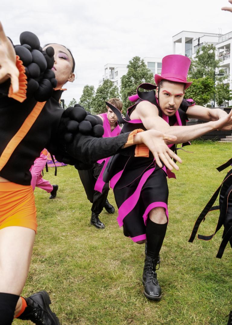 Dansare i svarta kläder med mycket orange och rosa detaljer dansar mot kameran.