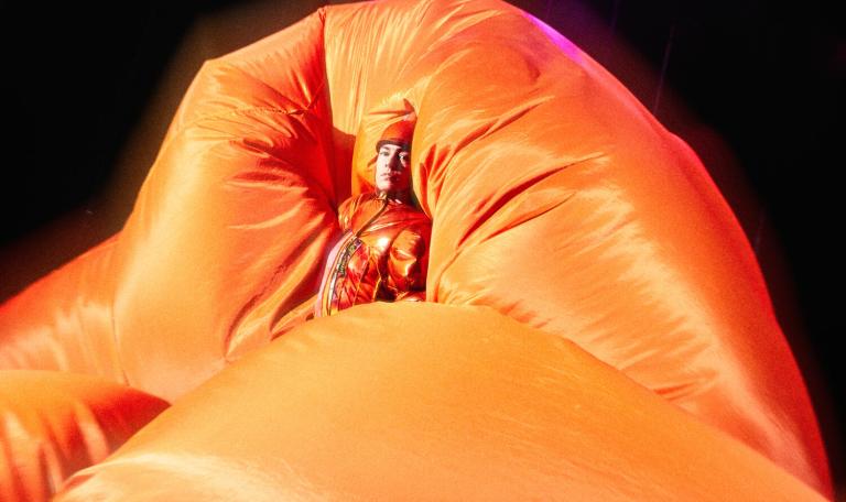 En dansare i orange kläder står omsluten av en orange balongliknande form i dansföreställningen SORRY NOT SORRY.
