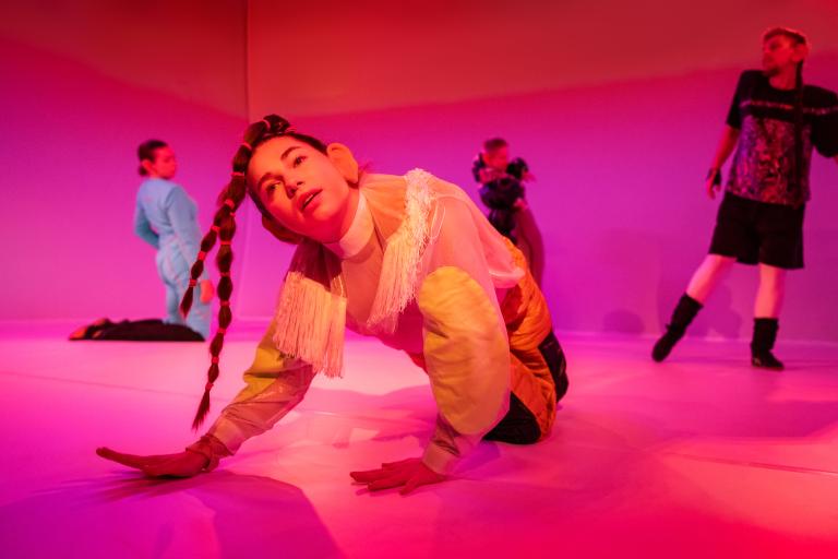 FRamför tre andra dansare lutar sig en dansare i glansiga ljusa kläder fram med händerna på golvet och tittar drömskt uppåt.