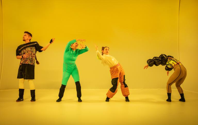 Fyra dansare dansar böjda åt olika håll med armarna pekande framåt och bakåt i ett gulfärgat rum.