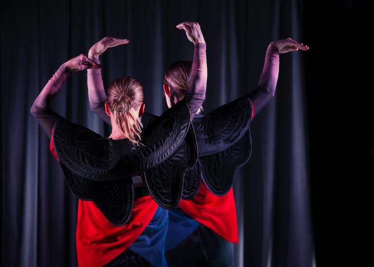 Två dansare står bakom varandra med ryggen mot dig och armarna uppe i luften och händerna hållna som näbbar.