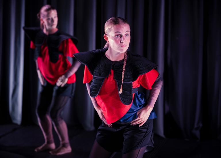 Två dansare i röda tröjor och svarta axlar med händerna på höfterna tittar över sin axel.
