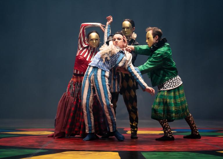 En dansare i blårandig kostym med kråsig skjorta lutar sig bakåt i armarna på tre dansare med guldmasker.