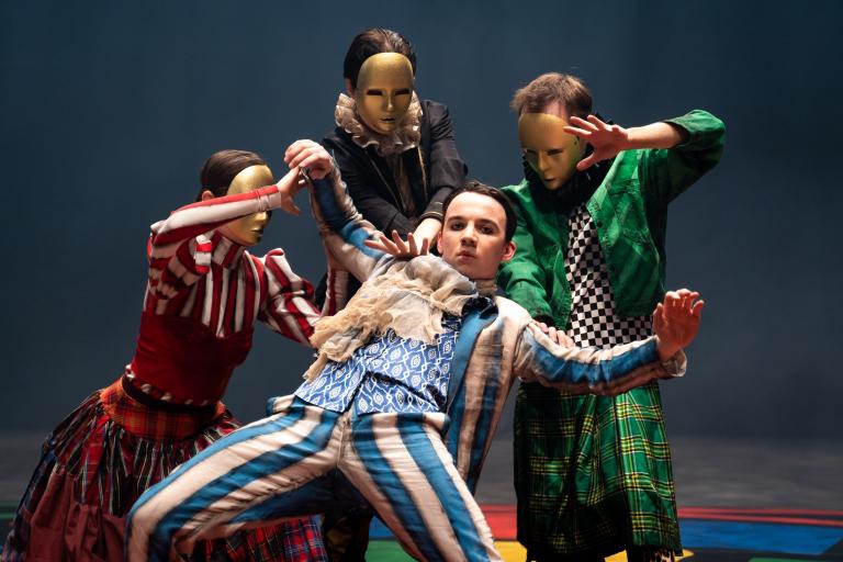 En dansare i blårandig kostym och kråsig skjorta lutar sig bakåt i armarna på tre dansare i guldmasker.