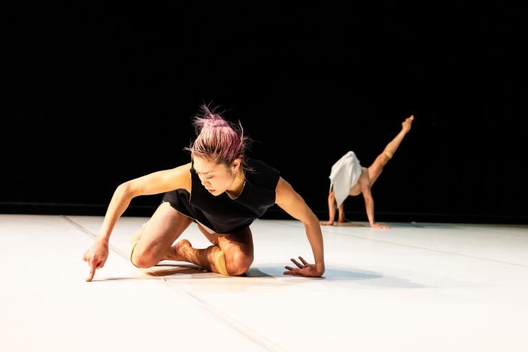 Dansaren Kat Nakui sitter på knä nära i bilden och trycker sitt pekfinger mot det vita golvet medan dansaren Samuel Denton står på händerna och ett ben med det andra benet spänt upp och utåt i luften i bakgrunden.