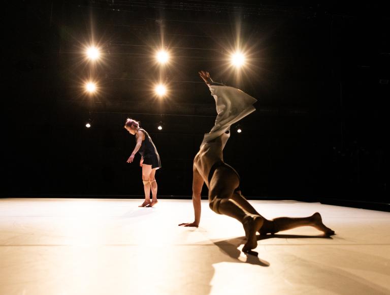 Dansaren Samuel Denton på händer och knän på ett vitt golv svänger upp sin arm med tyg fladdrande ut som en vinge. I bakgrunden står dansaren Kat Nauki i ljuset av strålkastare.