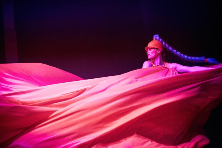 En dansare med en ridhjälmsliknande hatt där en lång blå fläta är fäst drar ett mycket stort tygstycke böljande genom luften i rosa ljus 