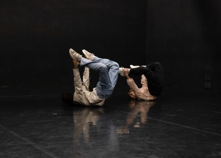 Ur dansföreställningen Megastructure, två dansare ligger på mage. De håller om sina fötter med benen böjda högt uppåt i luften.