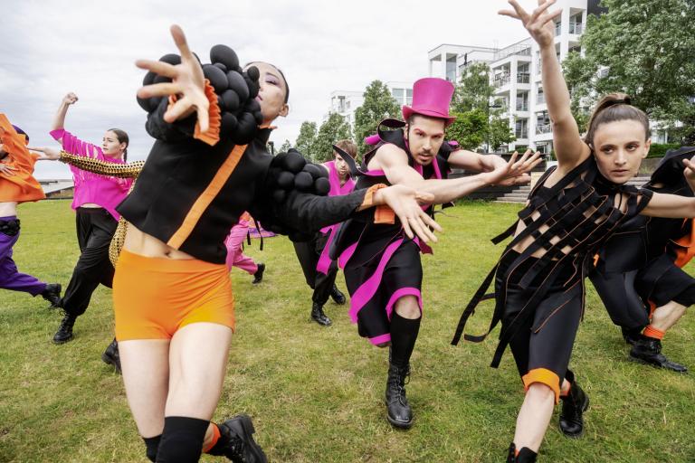 Dansare i svarta kläder med mycket orange och rosa detaljer dansar mot kameran.