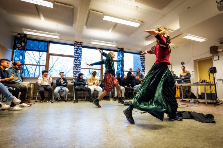 Två dansare i renässansliknande kläder i röd och grön sammet med gulddetaljer dansar med stora steg och utsträckta armar.