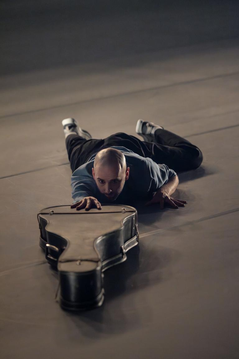 Dansaren Samuel kryper på magen på golvet och sträcker sin arm mot ett gitarrfordral i Skånes Dansteaters jubileumsföreställning Wing Ding av koreorgafen Lidia Wos.