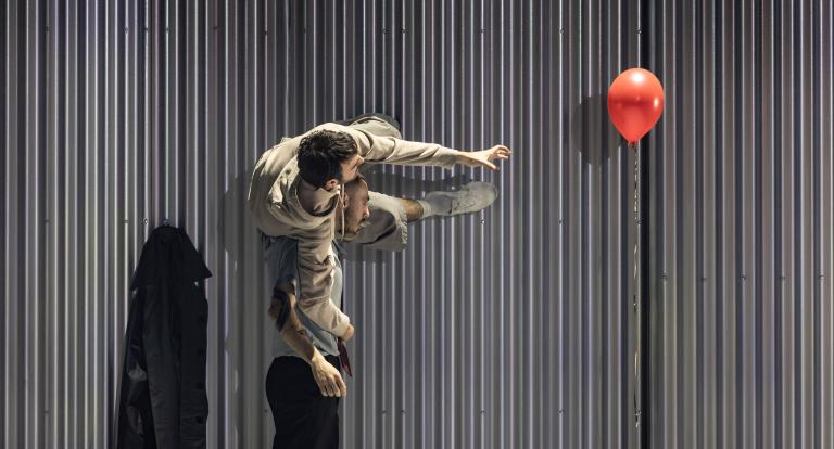Dansaren Freddy bär dansaren Facundo på sina axlar så att Facundo kan gå längs en vägg mot en röd balong i Skånes Dansteaters jubileumsföreställning Wing Ding av koreorgafen Lidia Wos.