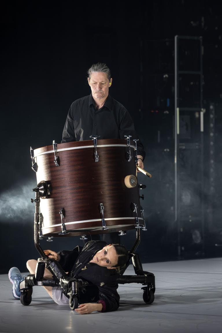 Dansaren Hanna ligger under en stor trumma på hjul som följs av en slagverkare i Skånes Dansteaters jubileumsföreställning Wing Ding av koreorgafen Lidia Wos.