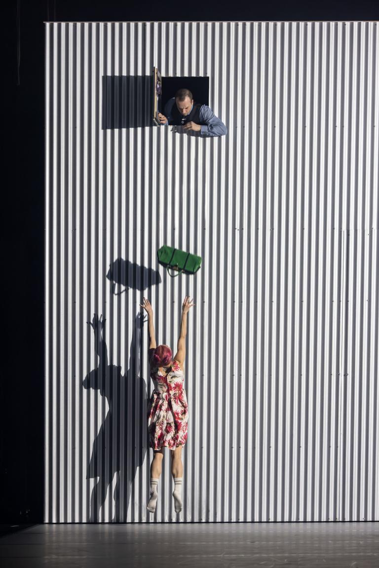 Dansaren Tomas släpper en grön handväska från en lucka högt upp på en vägg ner till dansaren Kat i röd- och vitblommig klänning som försöker fånga den i Skånes Dansteaters jubileumsföreställning Wing Ding av koreografen Lidia Wos.