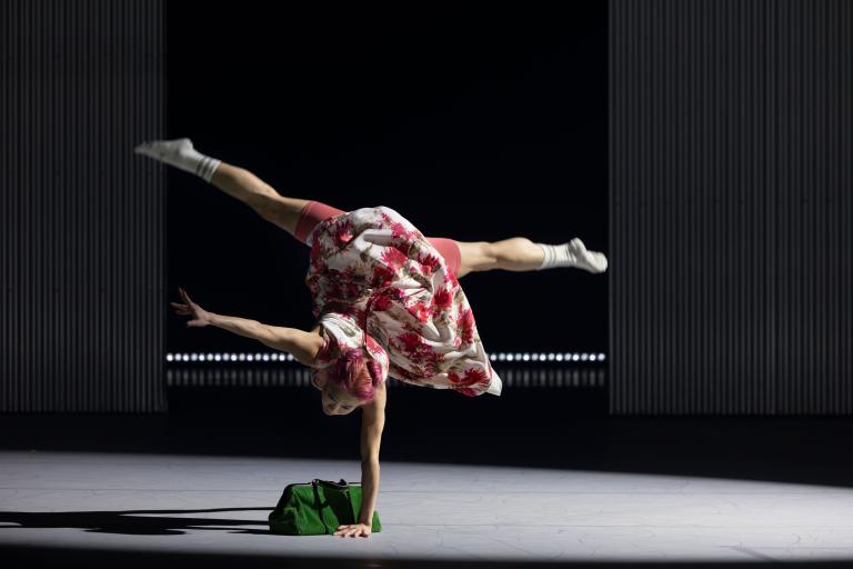 Dansaren Kat i röd- och vitblommig klänning står på en hand med benen i split vid en grön handväska i Skånes Dansteaters jubileumsföreställning Wing Ding av koreografen Lidia Wos. Foto: Tilo Stengel.