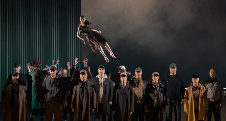 Dansaren Hanna i brun tyllkjol kastas svävande upp i luften ovanför huvudena på en grupp av Malmö OPEras körsångare klädda i mörka rockar och kepsar i Skånes Dansteaters jubileumsföreställning Wing Ding av koreorgafen Lidia Wos. Foto: Tilo Stengel.