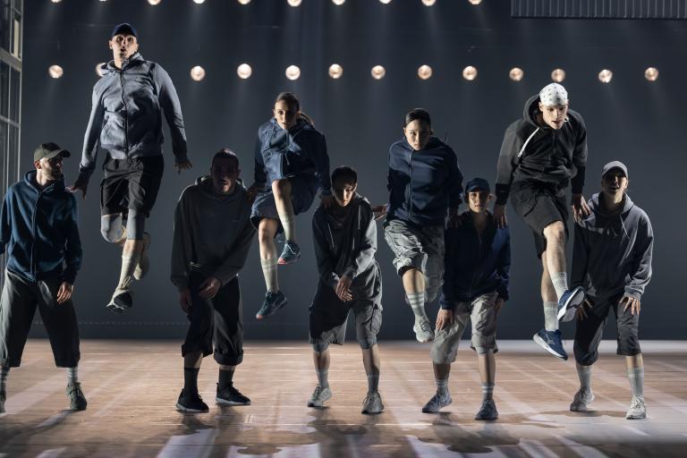 Nio dansare i hoodies, kepsar och shorts i gråblå nyanser står på rad. Varannan dansare studsar upp i luften lyft på axlarna av dansarna runtomkring i Skånes Dansteaters jubileumsföreställning Wing Ding av koreografen Lidia Wos. Foto: Tilo Stengel.