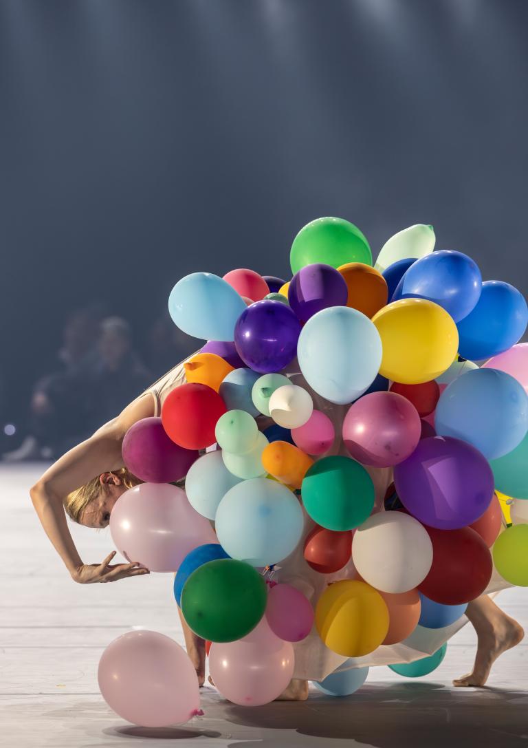 Dansaren Matilda klädd i en kjol täckt av balonger i regnbågens alla färger böjer sig på tå ner mot golvet med armen i en båge över huvudet i Skånes Dansteaters jubileumsföreställning Wing Ding av koreografen Lidia Wos. Foto: Tilo Stengel.