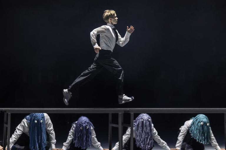 Dansaren Emma Klädd i svarta byxor och vit skjorta springer på ett långt bord. Under bordet sitter dansare klädda som Emma men med fransiga peruker i blålila toner med runda ögon på i Skånes Dansteaters jubileumsföreställning Wing Ding av koreorgafen Lidia Wos.