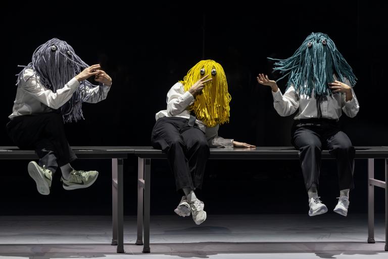 Tre dansare klädda i vita skjortor och svarta byxor med fransiga peruker som täcker deras ansikten sitter på bord och dinglar med benen i Skånes Dansteaters jubileumsföreställning Wing Ding av koreografen Lidia Wos. Foto: Tilo Stengel.