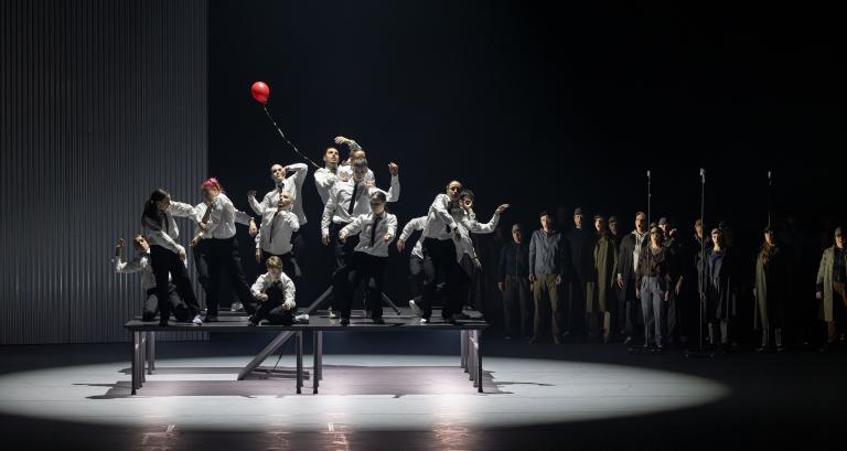 Fjorton dansare i vita skjortor och svarta byxor och slipsar dansar på ett stort bord med en liten röd balong. Bakom dem står en stor grupp sångare ur Malmö Operas kör.