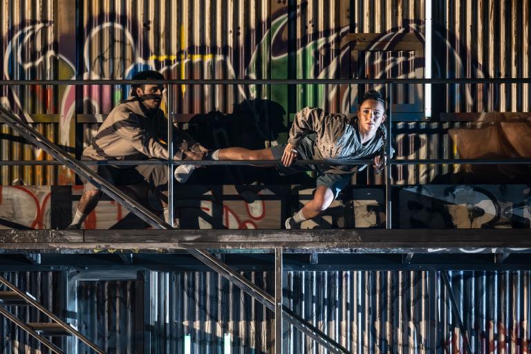 Dansaren Velntina står med ett ben på relingen och andra på en avsats. Dansaren Facundo håller i hennes ena fot. Bakom dem en veckad plåtvägg med graffitti.