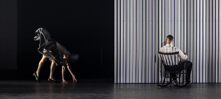 Dansaren Freddy sitter i en svart gungstol vid en vägg med ryggen mot dig. Han tittar snett över axeln mot en dansare i svart yvig tyllkjol som går på alla fyra på sträckta ben och armar i Skånes Dansteaters jubileumsföreställning Wing Ding av koreografen Lidia Wos.