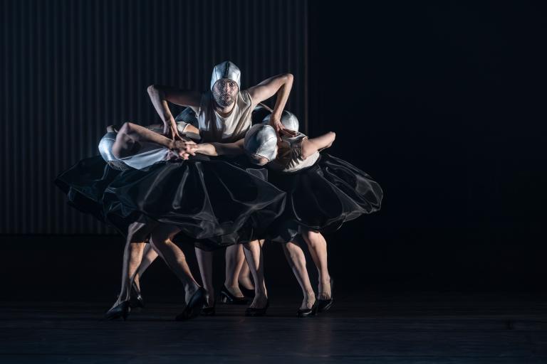 DAnsaren Sam stor omringade av dansare som cirkulerar nedåtböjda runt honom som en kjol. De har alla stela svarta tyllkjolar, högklackade skor och en silvrig tunn hjälmliknande mössa med silvrigt flor.