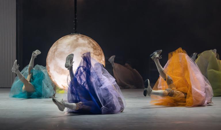 Dansare i färgglada fluffiga tyllkjolar ligger på rygg och sträcker benen upp i luften. Mitt bland dansarna hänger ett månliknande stort klot som lyser.