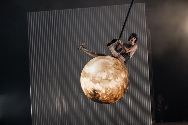 Dansaren Carl sitter och gungar på en rivningskula som ser ut som en fullmåne.  Kulan svingar genom luften och CArl i discobollshjälm sträcker ut benen för att ta mer fart i Skånes DAansteaters jubileumsföreställning Wing Ding av koreografen Lidia Wos.