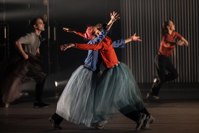 I förgrunden står dansarna Kat och Emma lutade mot varandra som i en kram, men deras armar stretar ut bakom den andre. De har ljusblå tyllkjolar och skjortor i klara färger.
