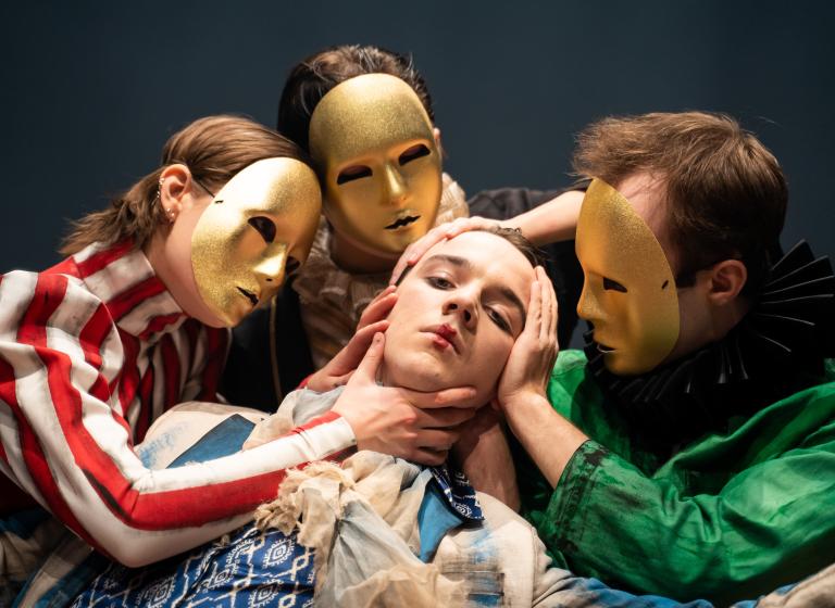 Tre dansare med guldmasker för sina ansikten håller sina händer runt ansiktet på en annan dansare.