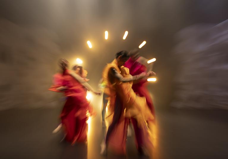 Dansare i fladdriga korallfärgade kläder dansar böljande framför en ring av ljuspunkter.