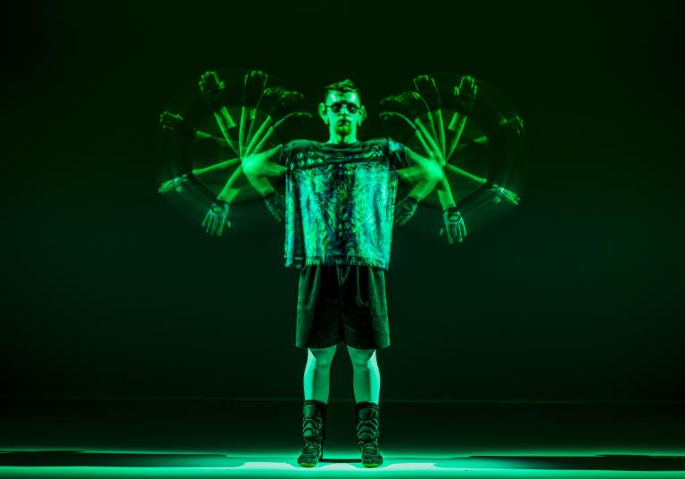 En dansare i mörk t-shirt, shorts och solglasögon står i grönt ljus och snurrar på armarna som propellrar. 