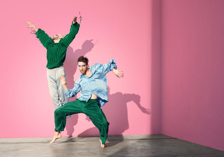 Två dansare i ljusblå och gröna fladdriga kläder dansar framför en rosa vägg.