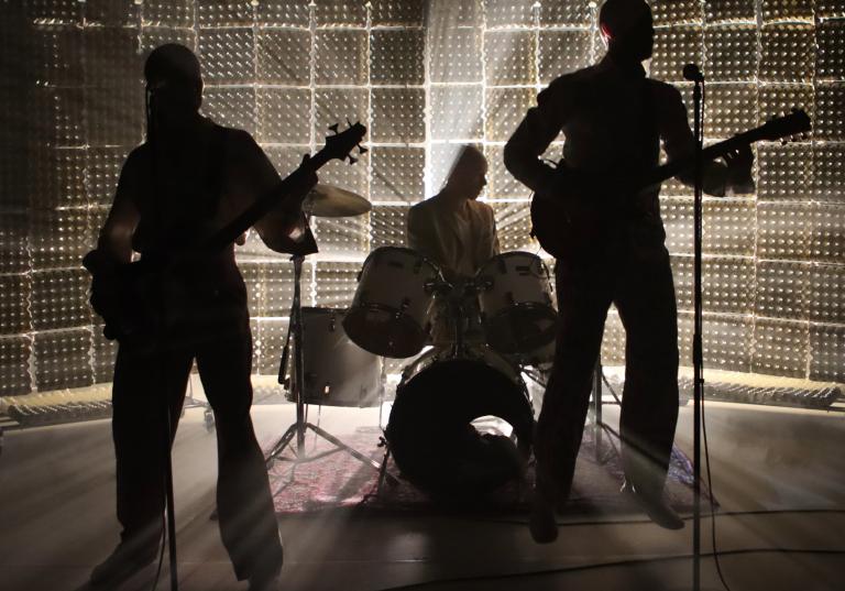 Tre personer med trummor, bas och elgitarr står på en scen i motljus så att vi endast ser deras siluetter och ljuset som strålar runt dom.
