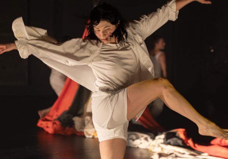 Dansaren Hazuki Kojima kädd i vita shorts och skjorta sträcker ut sina armar som vingar med skjortan fladdrande runt henne i Skånes Dansteaters dansföreställning Attempting Alchemy av koreografen Ludvig Daae.