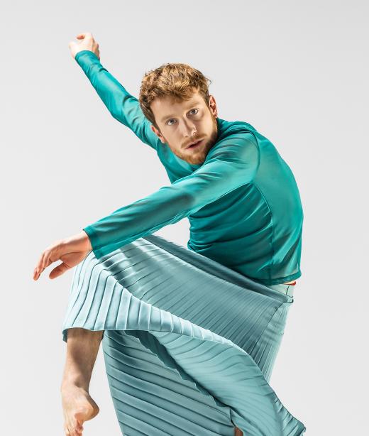 Bild på dansaren Jure Gostincar i turkos tröja och kjol i rörelse. 